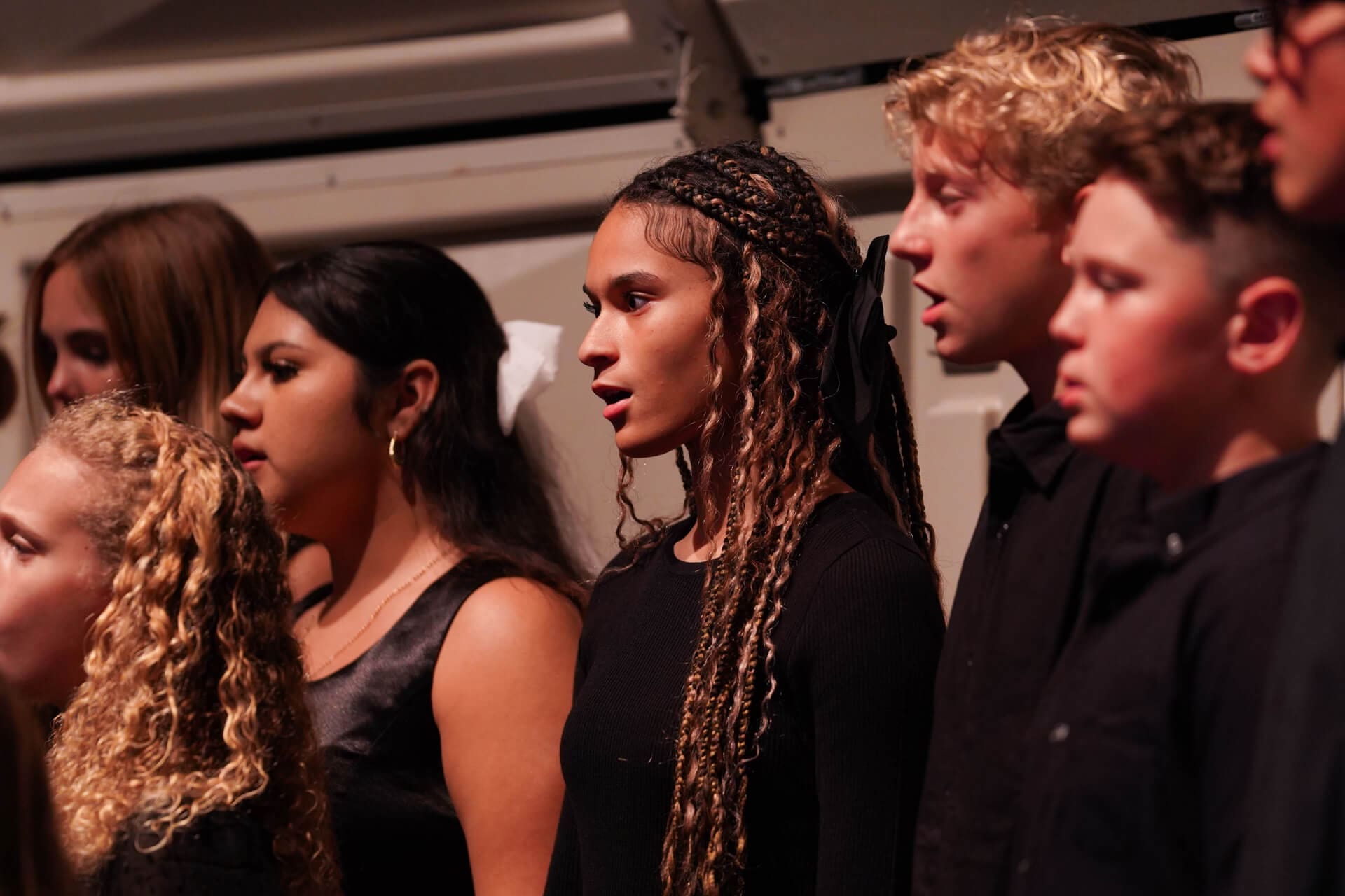 A group of young people, dressed in black, singing together in a choir. They stand in rows and appear focused, with their mouths open mid-song against a plain indoor background.