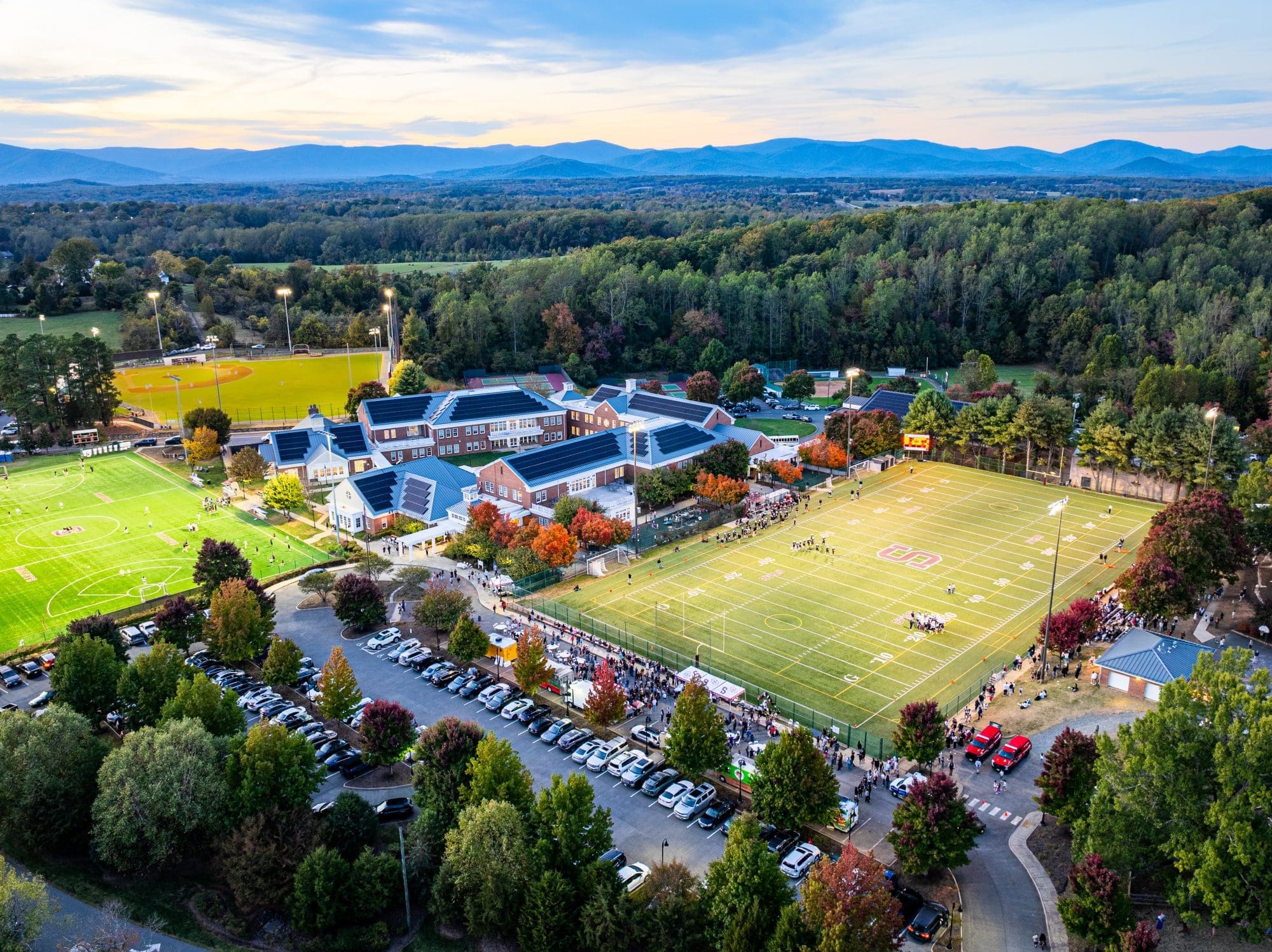Aerial view of a school campus with two sports fields, parking lot full of cars, and wooded hills in the background at sunset. Football game is in progress on the right field.