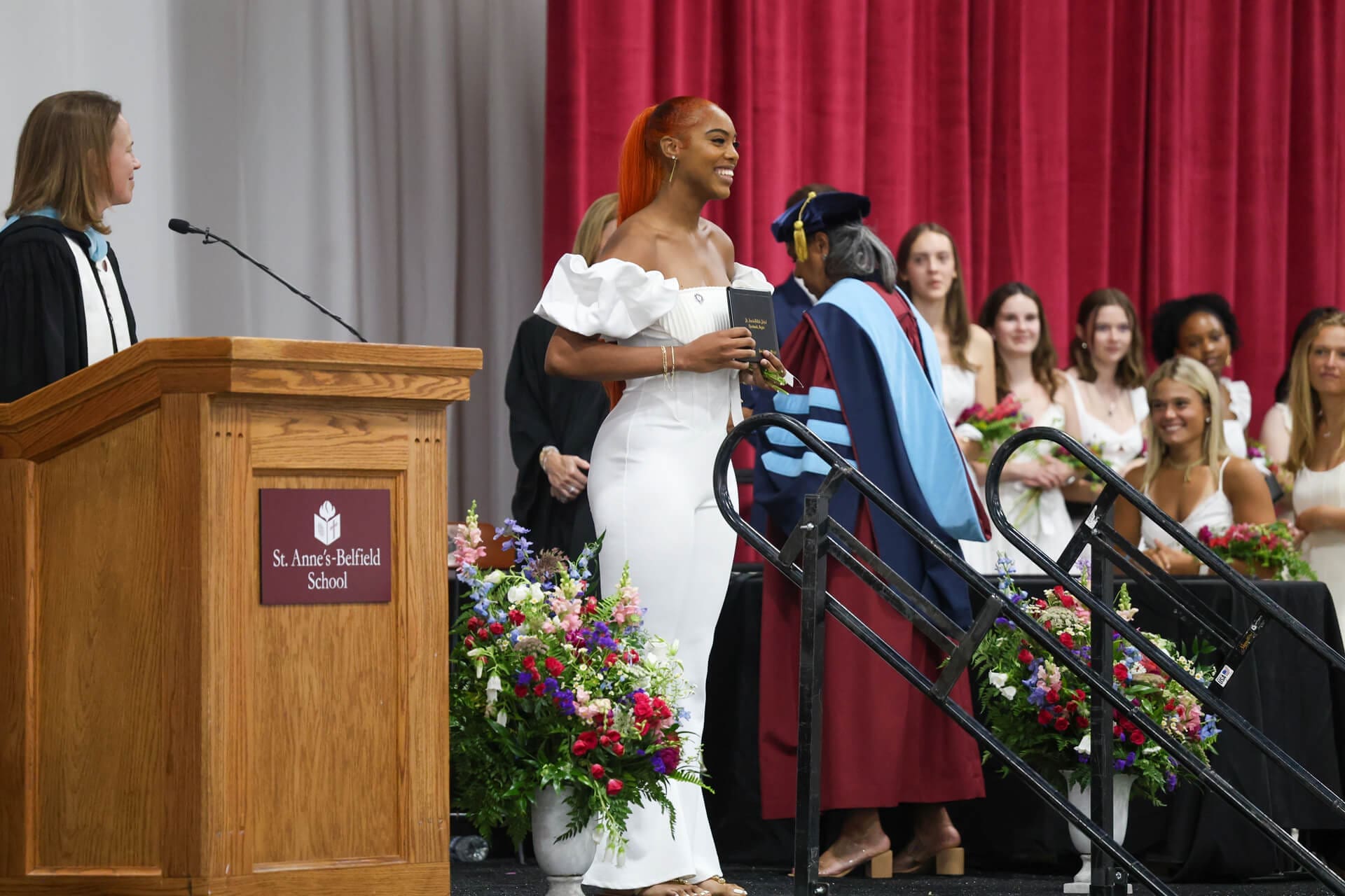 A graduate in a white dress smiles while receiving her diploma on stage at a ceremony. Other graduates in white stand nearby, and a woman at a podium watches. The stage is decorated with flowers and red curtains.