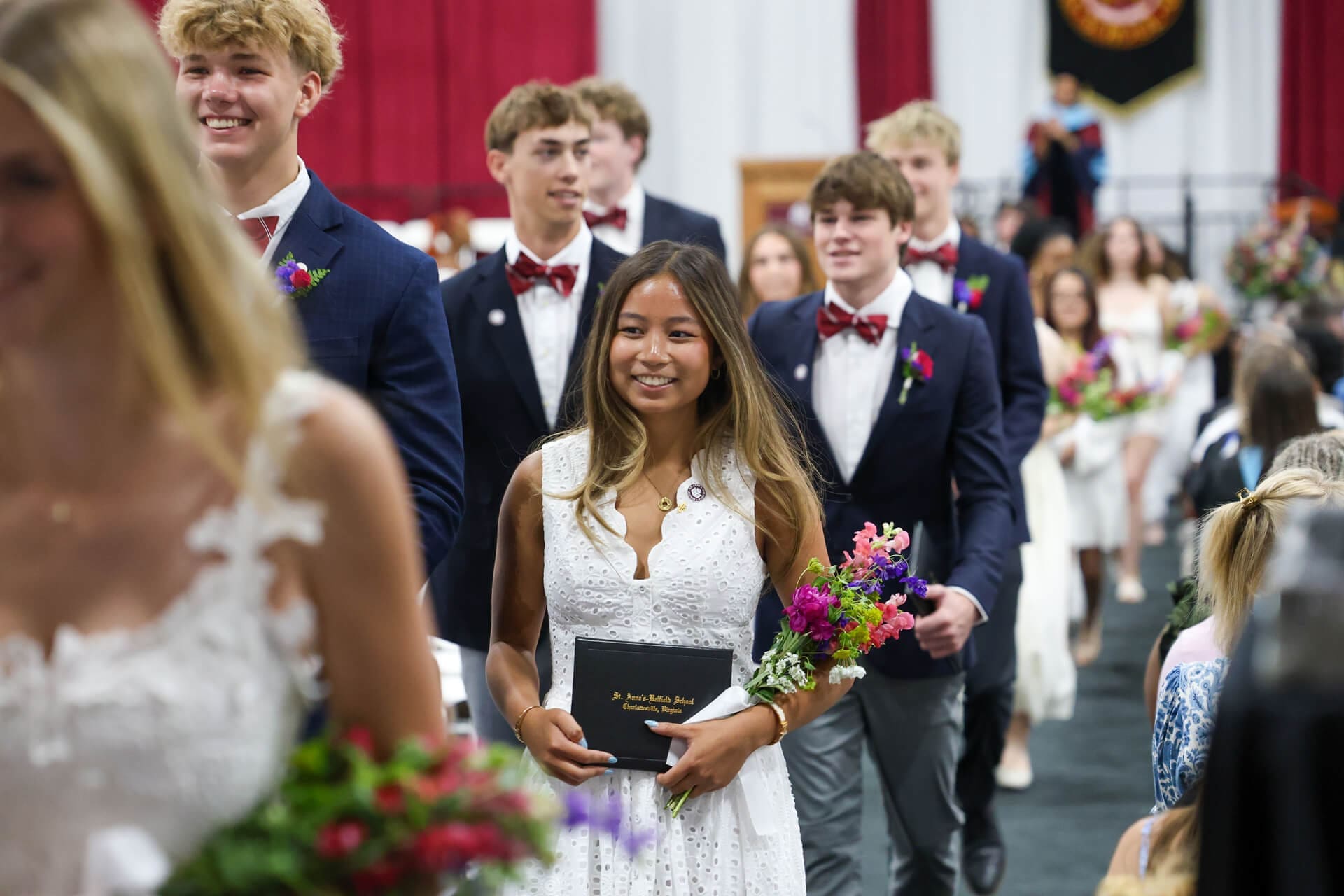 A young woman in a white dress holds a diploma and flowers, smiling as she walks in a procession with other graduates in formal attire during a graduation ceremony.