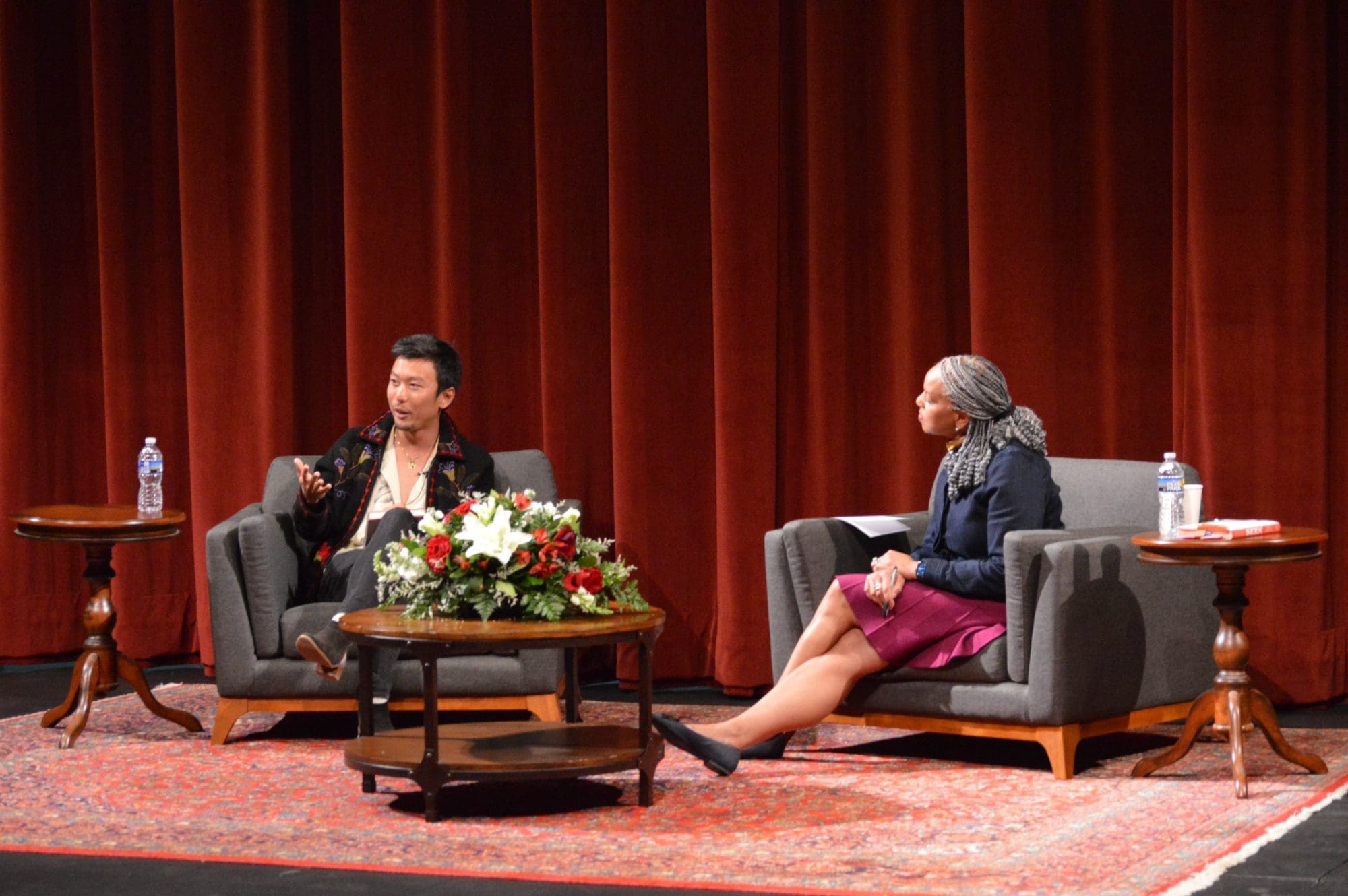 A man and a woman seated in armchairs on a stage participate in an Inspiration Speaks interview in front of a red curtain.