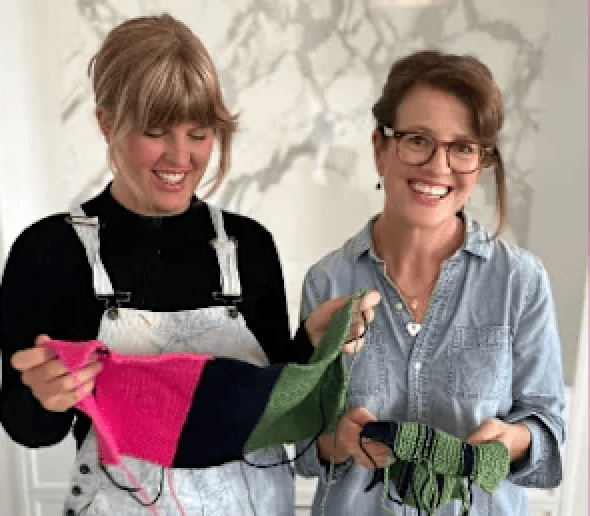 Two smiling women holding up knitted fabric projects.