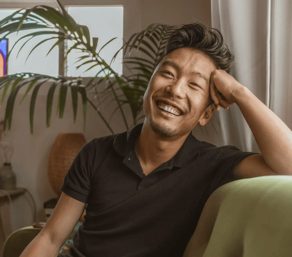A smiling man sits on a couch with his hand resting against his head.
