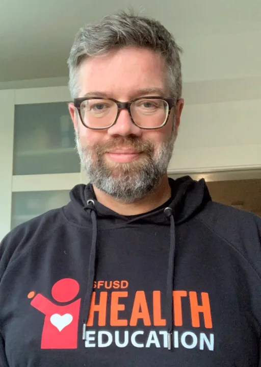 A man with graying hair and a beard wears glasses and a black SFUSD Health Education hoodie.