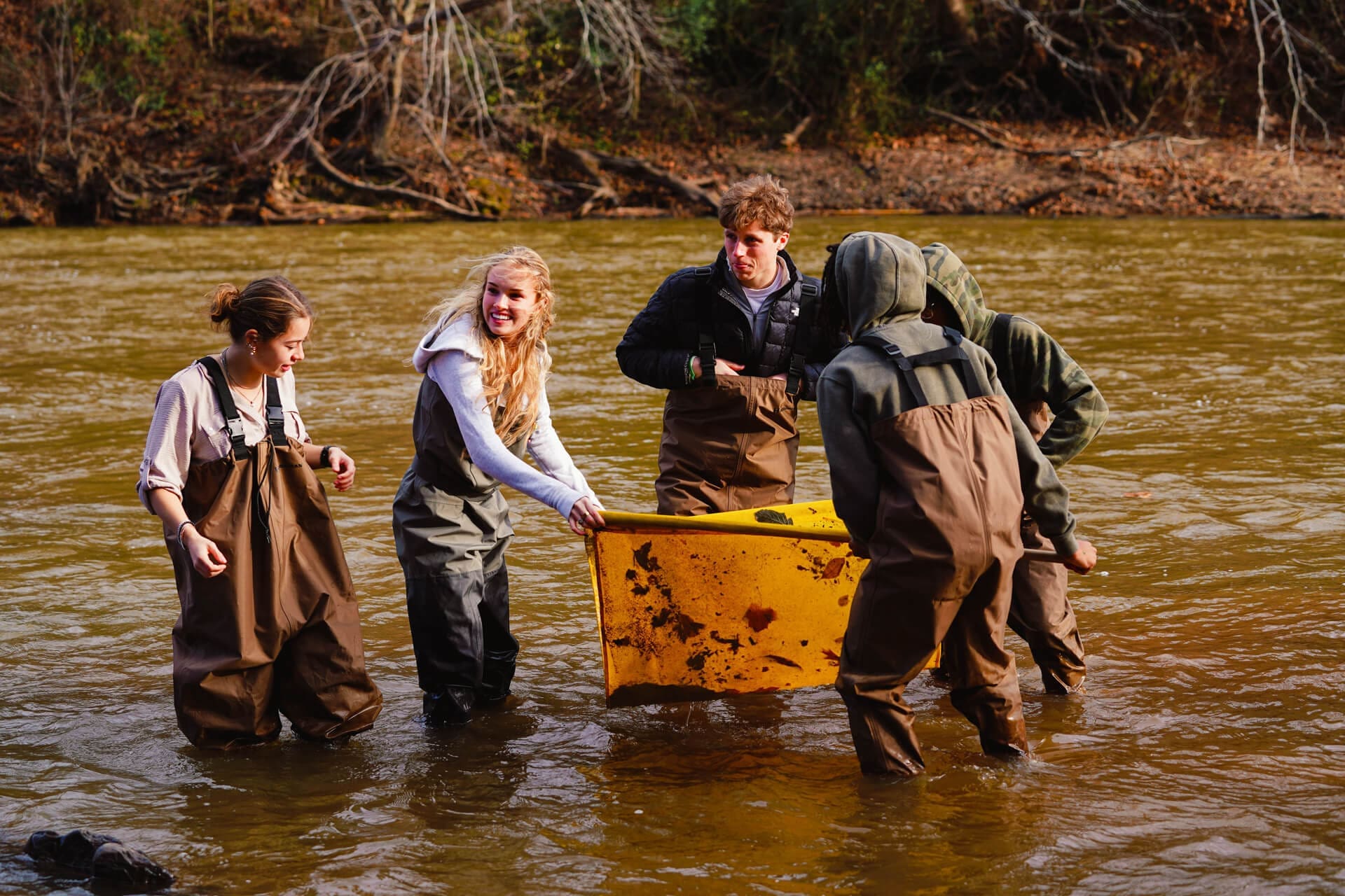 Five people wearing waders stand in a shallow river, likely as part of a school fieldwork project, holding a large yellow container. The group appears to be collecting samples, surrounded by trees and autumn foliage.