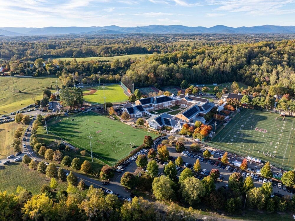 Aerial view of a school campus featuring multiple sports fields, parking lots, and lush trees, all set against rolling hills and mountains beneath a partly cloudy sky.