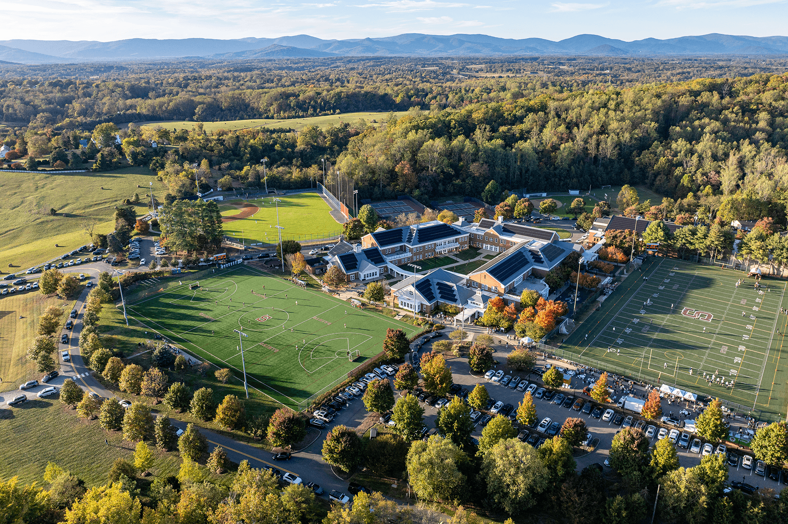 Aerial view of a school campus with sports fields, parking lots, and surrounding trees, set against a backdrop of rolling hills and distant mountains under a clear sky.