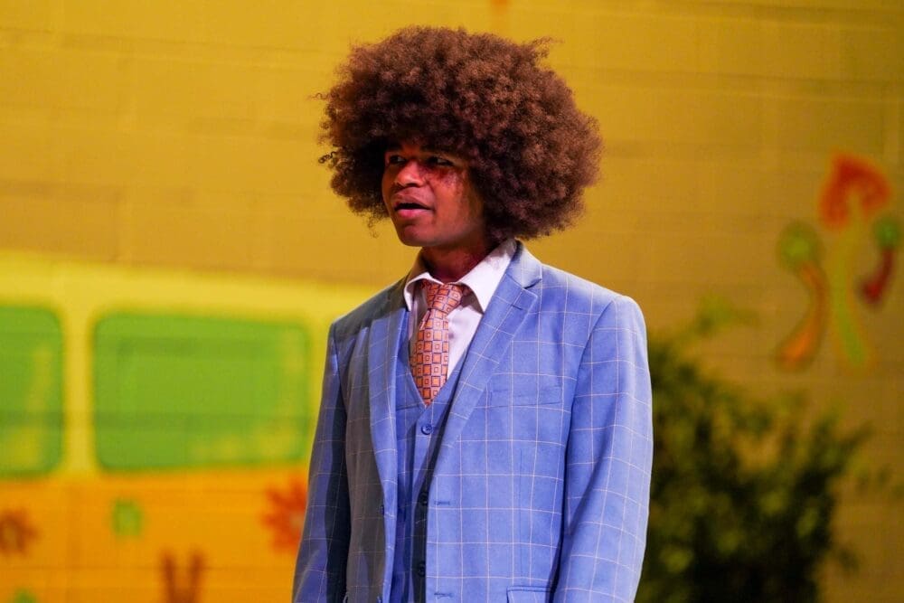 A person with a large afro hairstyle wears a light blue plaid suit, white shirt, and patterned tie, standing in front of a yellow and green mural with plants and abstract shapes near a school.