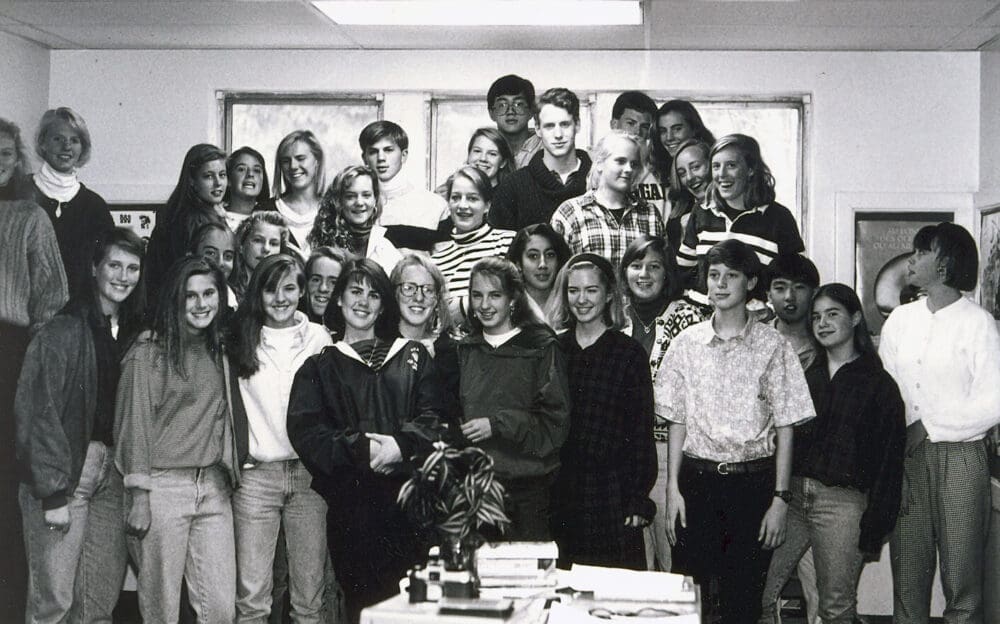 Black and white photo of a group of around 30 teenagers and a few adults standing close together indoors, smiling at the camera. Some are wearing casual clothes; there are windows in the background.