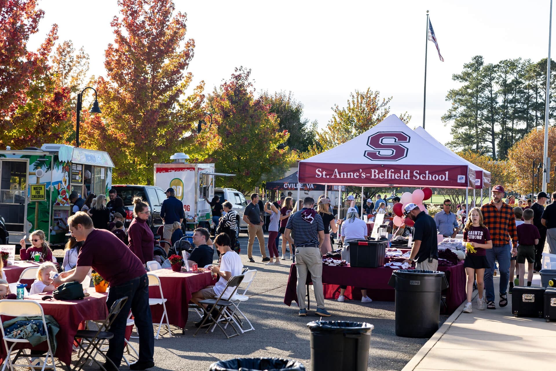 People gather outdoors at a school event with tables, food trucks, and a St. Anne’s-Belfield School tent. Trees with autumn leaves are in the background, and attendees are socializing and eating.