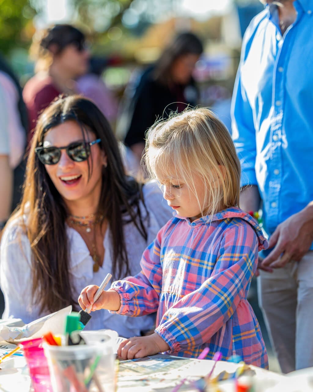 A young girl in a plaid dress colors at a table outdoors, while a smiling woman in sunglasses and a white shirt sits beside her. Other people are blurred in the background, enjoying a sunny day.