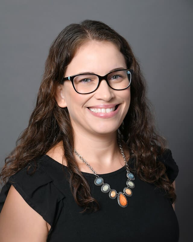 A woman with long brown wavy hair and glasses smiles at the camera. She is wearing a black top and a statement necklace with large oval stones, against a plain gray background.