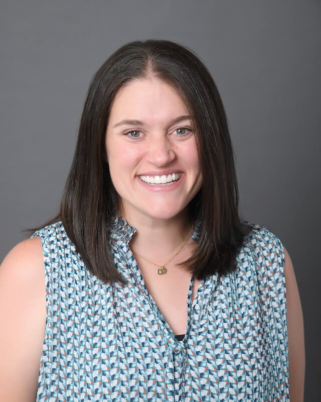 A woman with straight, shoulder-length brown hair smiles at the camera. She is wearing a sleeveless, patterned blouse and a small necklace. The background is plain gray.