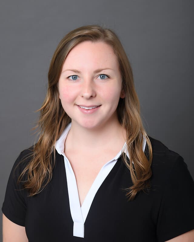 A woman with light brown hair wearing a black top with a white collar smiles at the camera against a plain gray background.