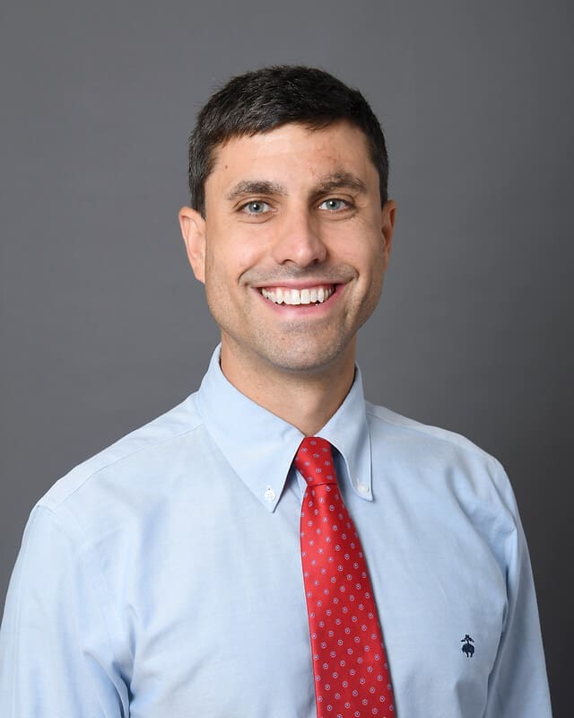 A man with short dark hair, wearing a light blue button-down shirt and a red tie with small white patterns, smiles at the camera against a plain gray background.