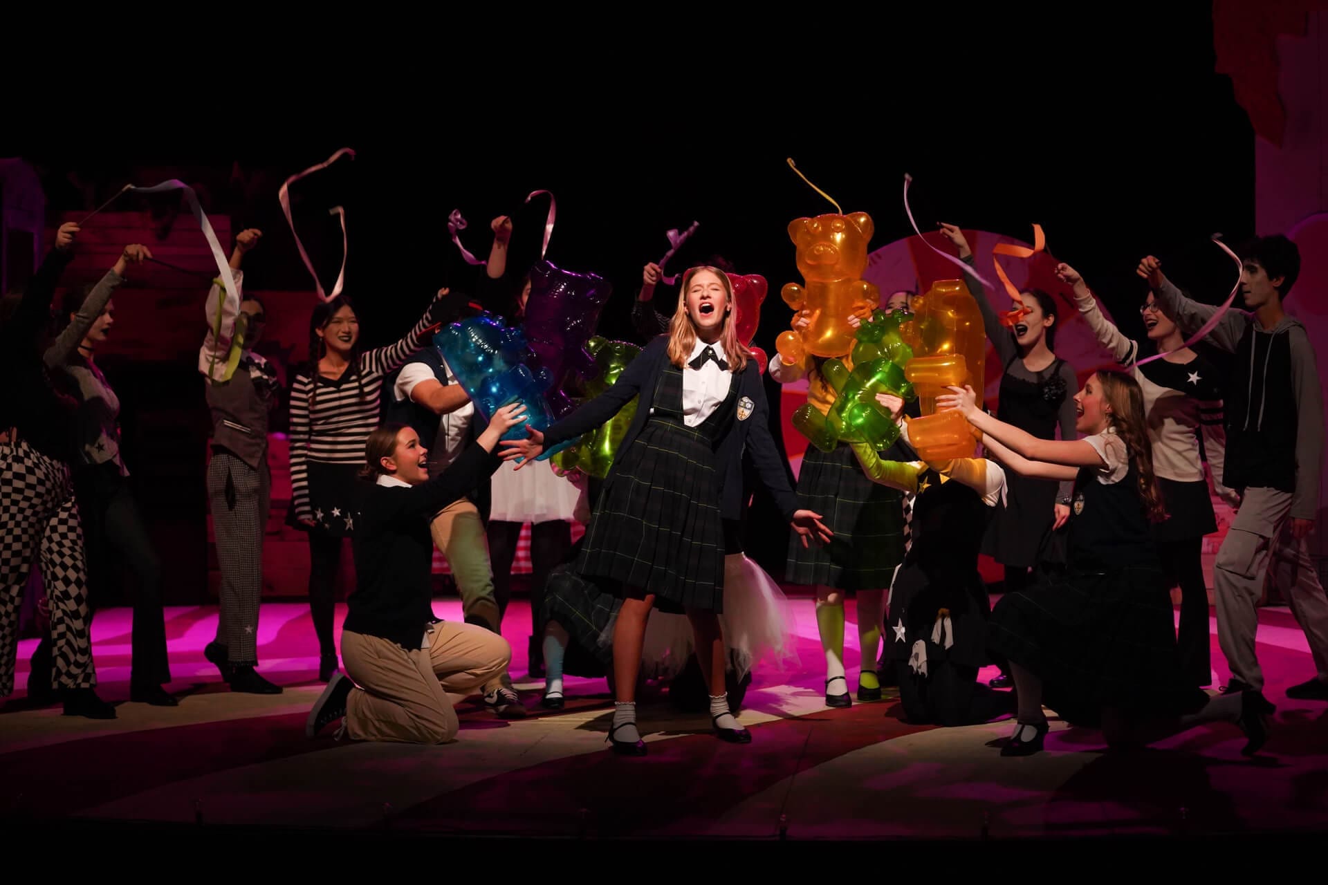 A group of young actors in school uniforms perform energetically on stage, holding large colorful gummy bear props, with one girl singing passionately at center stage under bright lights.