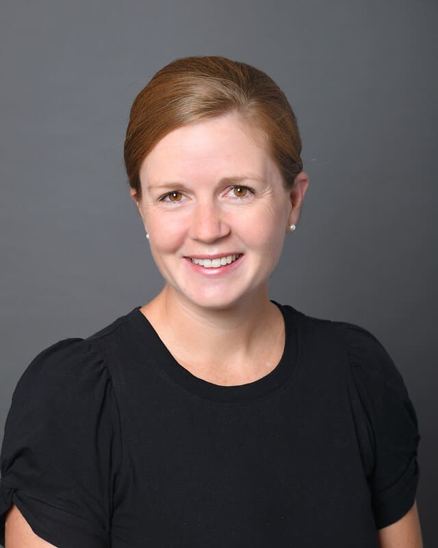A woman with light skin and brown hair pulled back, wearing a black top and small stud earrings, smiles in front of a plain gray background.