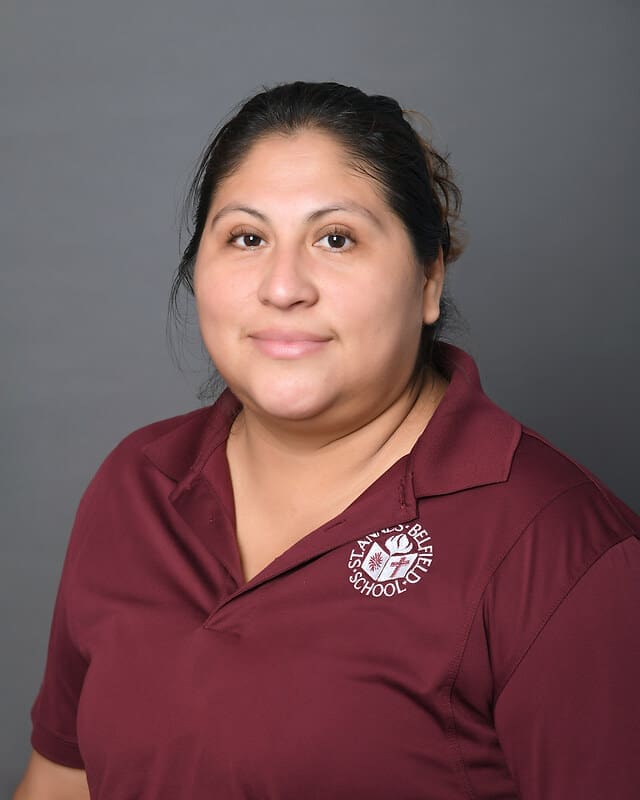 A woman with long dark hair pulled back, wearing a maroon polo shirt with a “La Mesa Elementary School” logo, poses and smiles in front of a plain gray background.