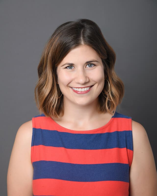 A woman with shoulder-length brown hair wearing a sleeveless red and navy blue striped top smiles in front of a plain gray background.