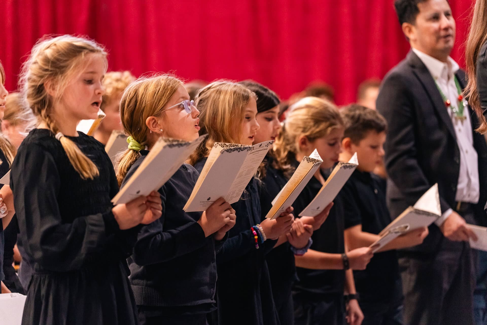 A group of children dressed in dark clothing sing from sheet music, standing in rows with a red curtain in the background. An adult in formal attire stands among them.