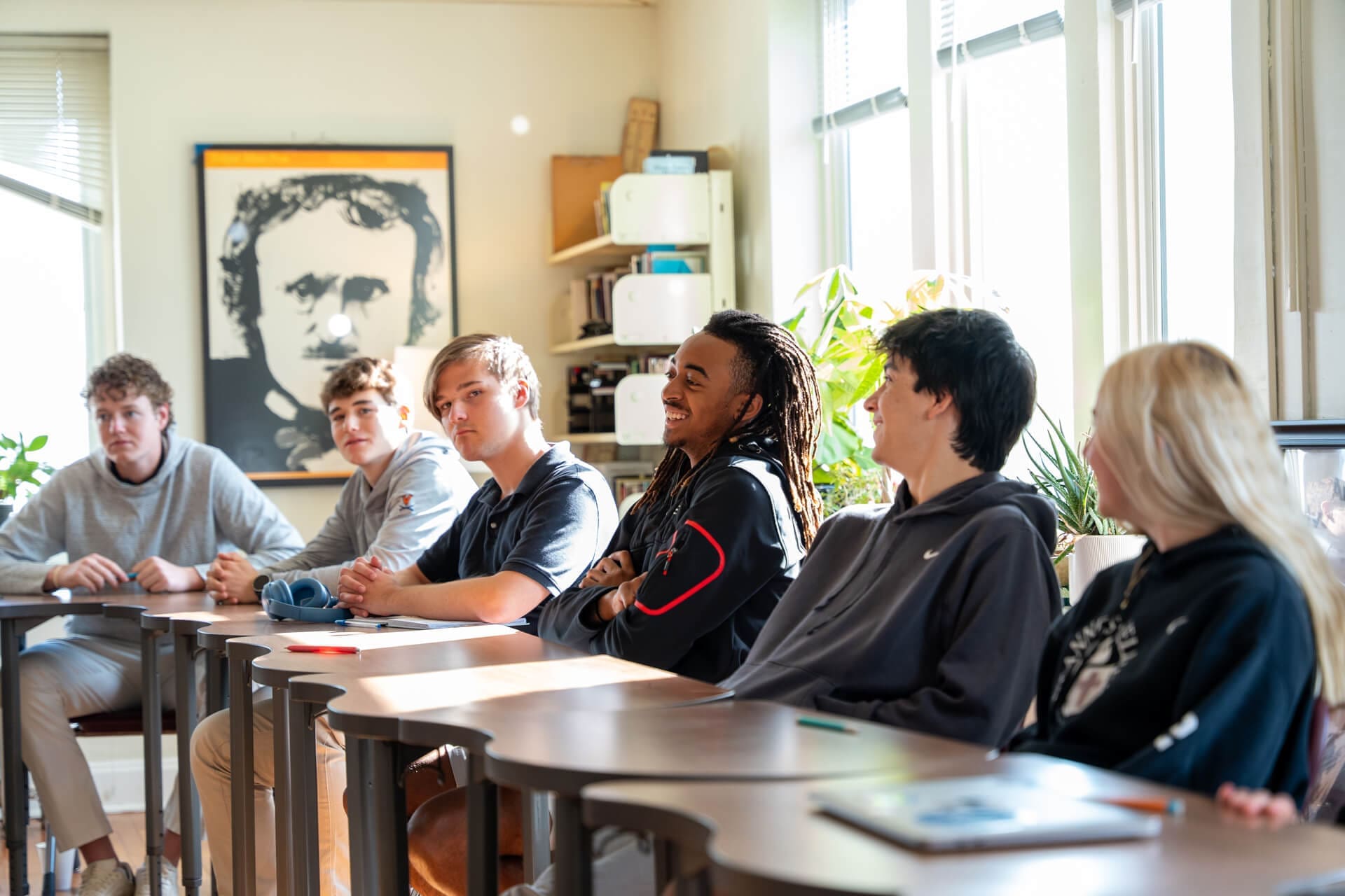 Five students sit in a brightly lit classroom, engaged in conversation. A large portrait hangs on the wall behind them. Sunlight streams through the windows, and plants are visible by the window.