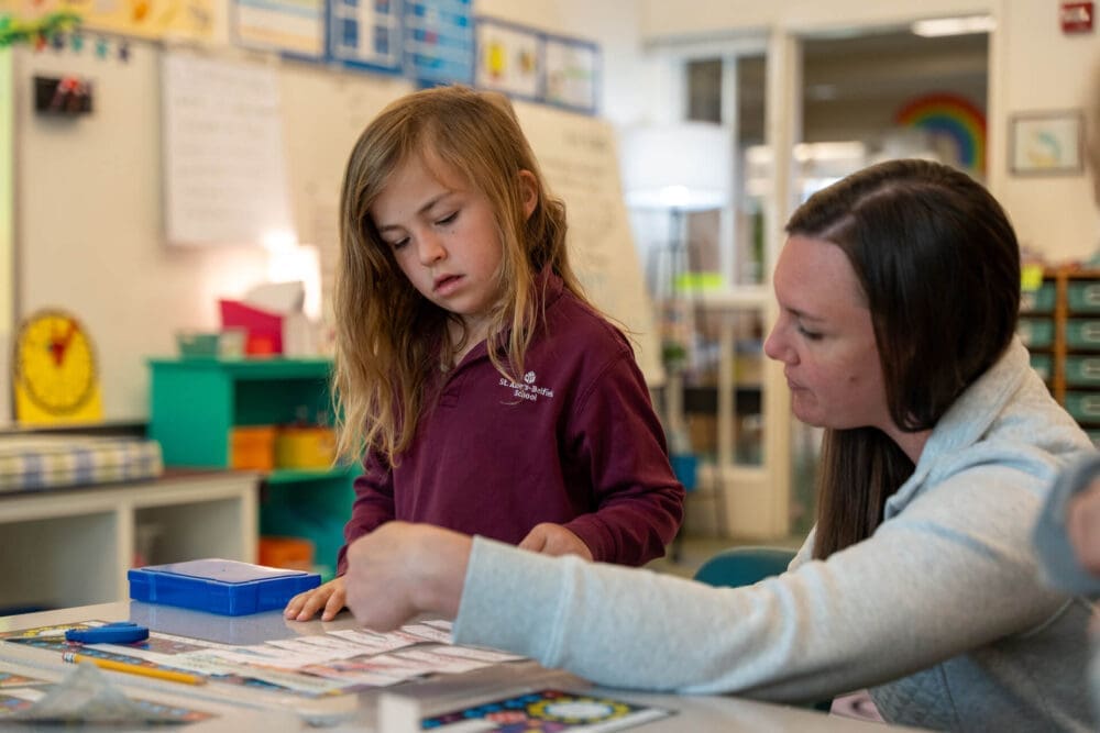 A young girl stands at a classroom table, looking at her work as an adult woman next to her helps point to something on a worksheet. The classroom is colorful and organized with shelves, posters, and educational materials.