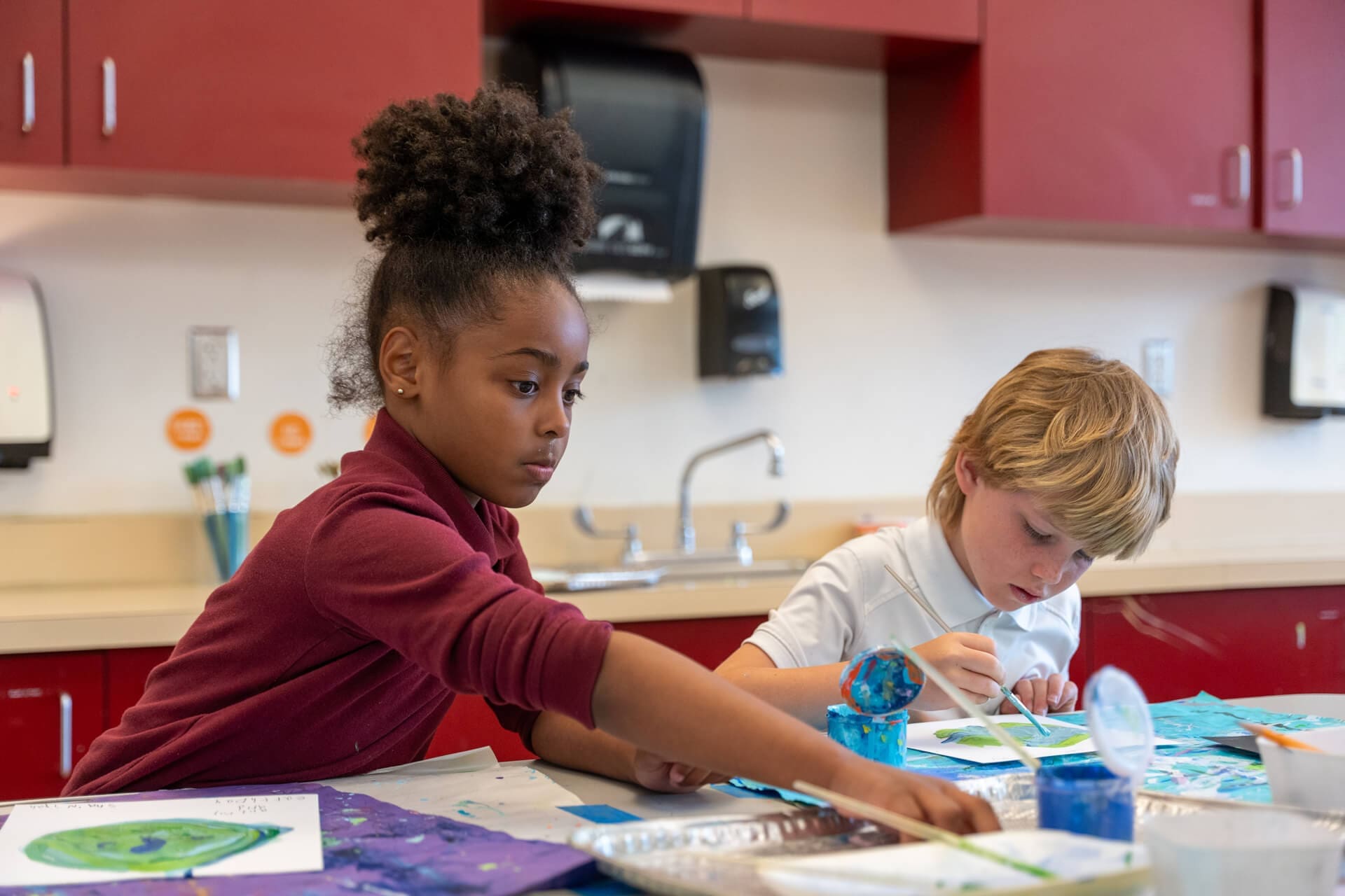 Two children sit at a table in a classroom, focused on painting with brushes and blue paint. The background shows red cabinets, a sink, and art supplies.
