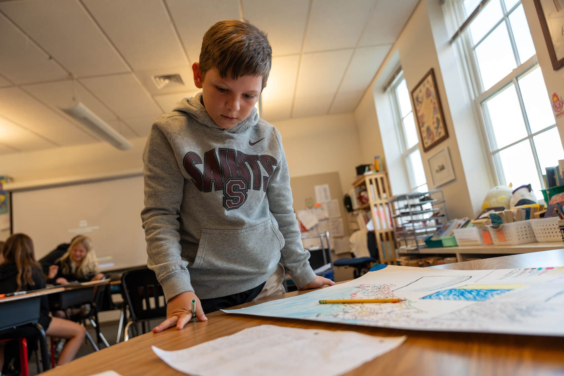 A boy in a gray hoodie stands at a school desk, looking at colorful drawings and papers. Other students are seated in the background, and sunlight streams through the windows.