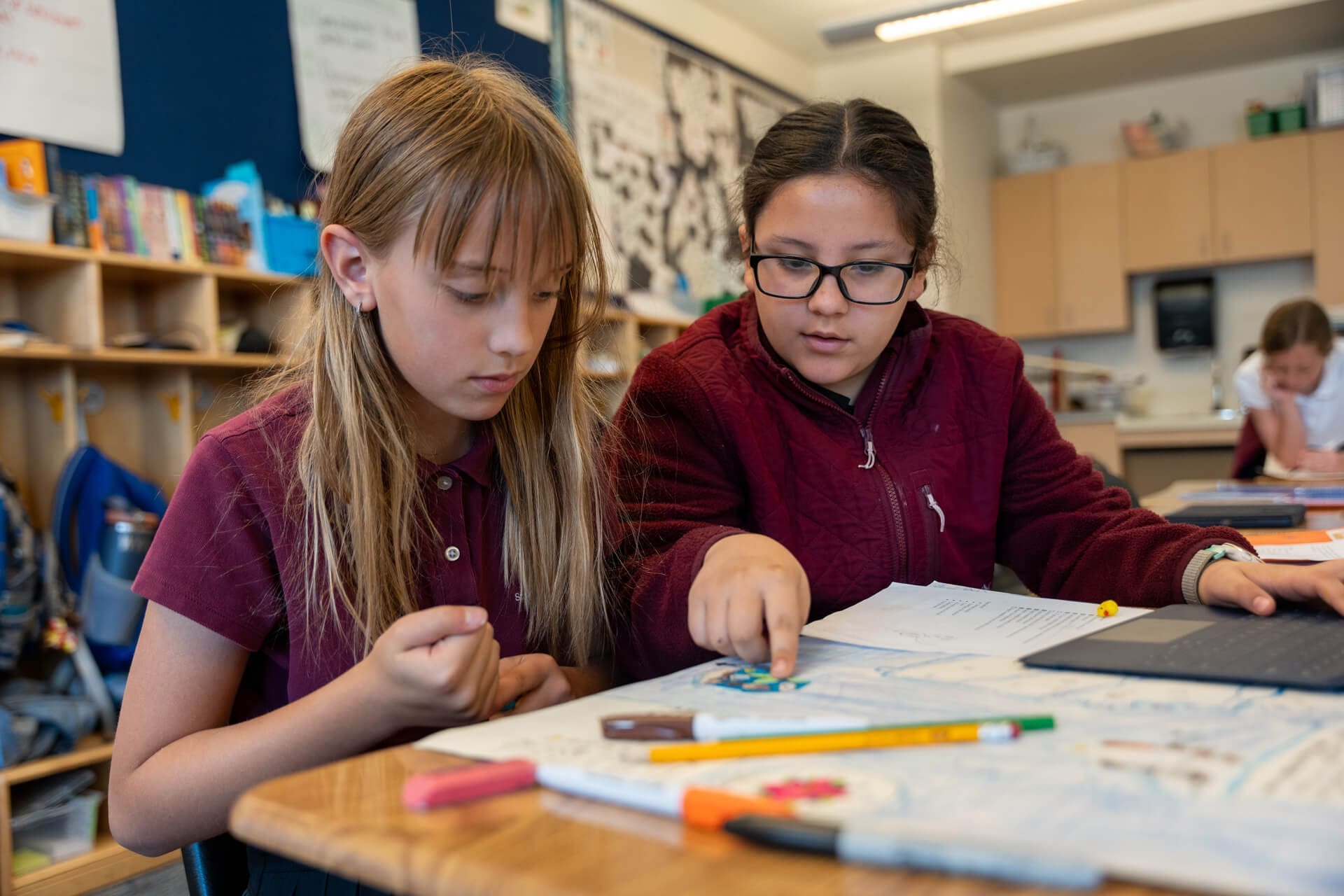 Two young students sit at a classroom desk, working together on a project. One points at a paper while the other looks on attentively. School supplies and papers are spread out in front of them.