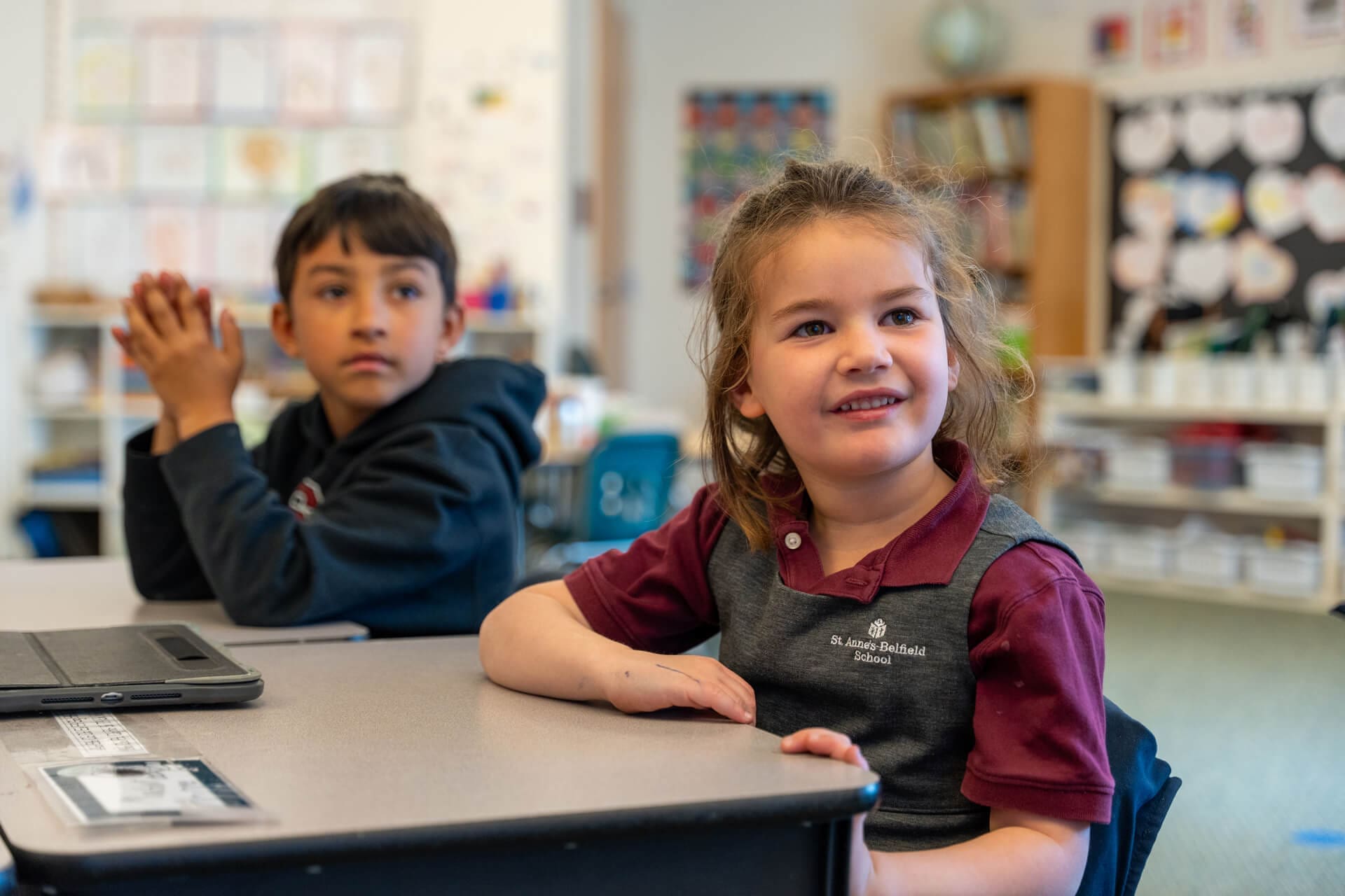 Two young children sit at desks in a colorful classroom, looking forward and smiling. The girl in front wears a maroon and gray school uniform shirt, and the boy behind her has a black hoodie. Shelves and artwork are visible in the background.