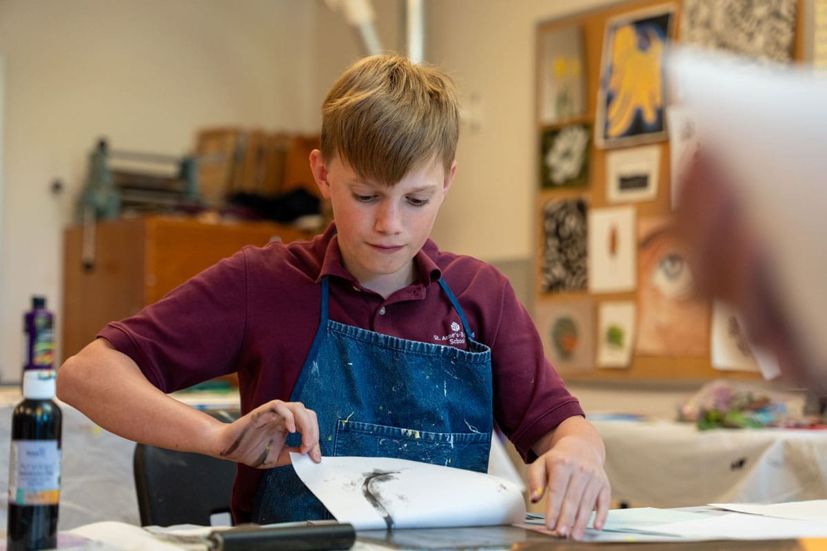 A boy wearing a maroon shirt and denim apron lifts paper from a desk, revealing a black and white print. Art supplies and drawings are visible in the background.