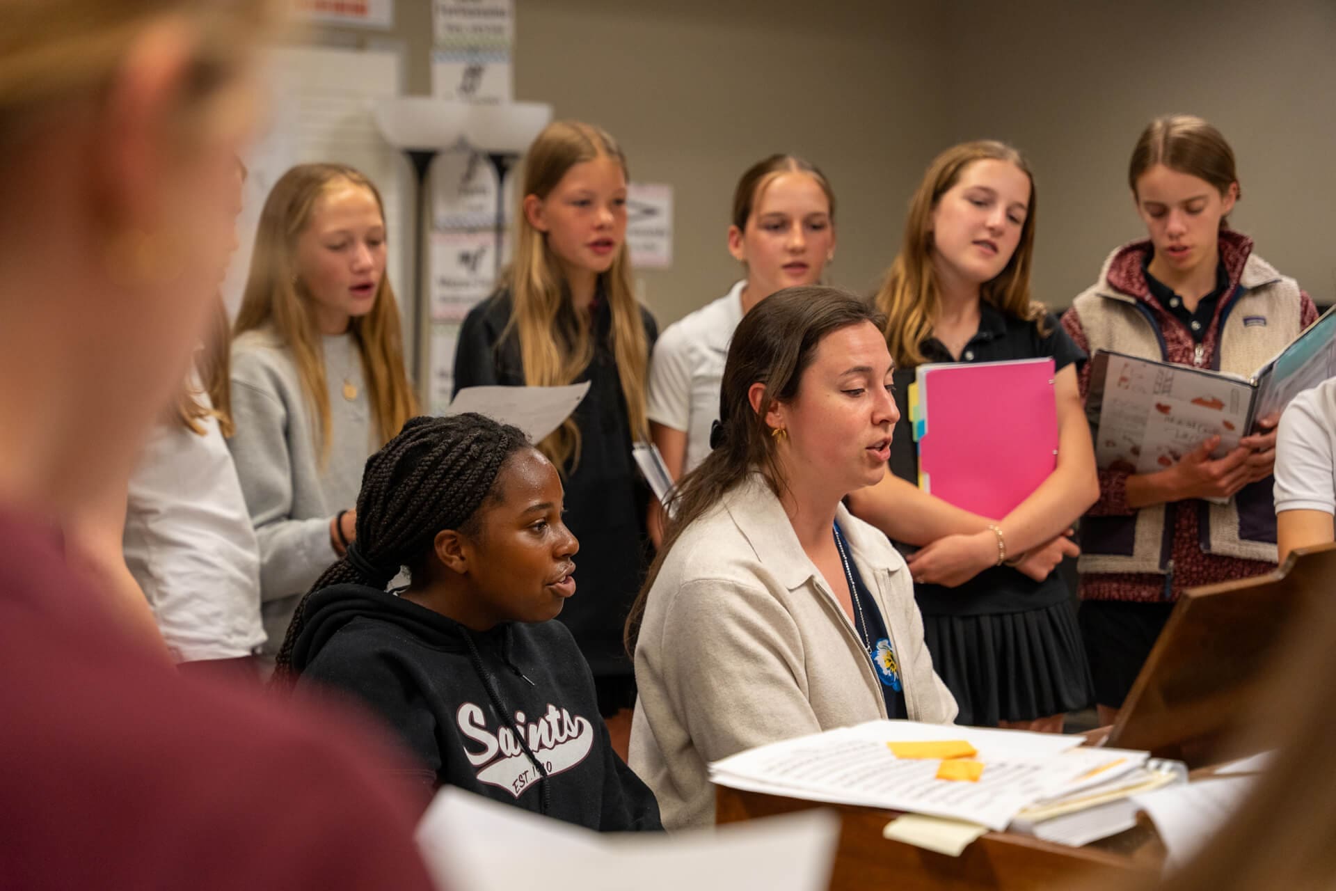 A group of girls and a teacher sit and stand around a piano, singing together in a classroom. Some hold folders or sheet music, and music posters are visible on the wall in the background.