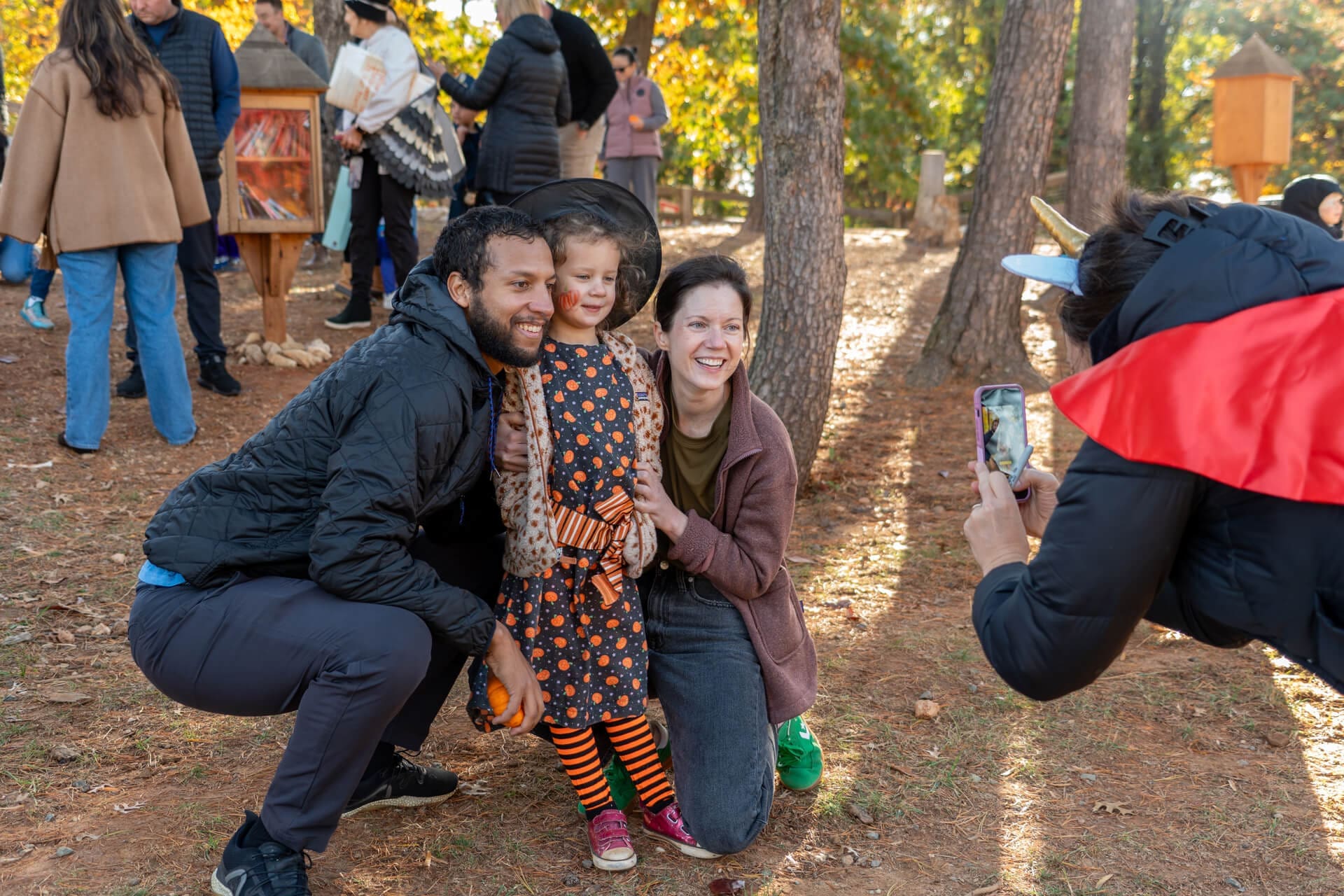 A young girl in a Halloween dress poses with two adults, all smiling for a photo being taken outdoors in a wooded area with people and trees in the background.