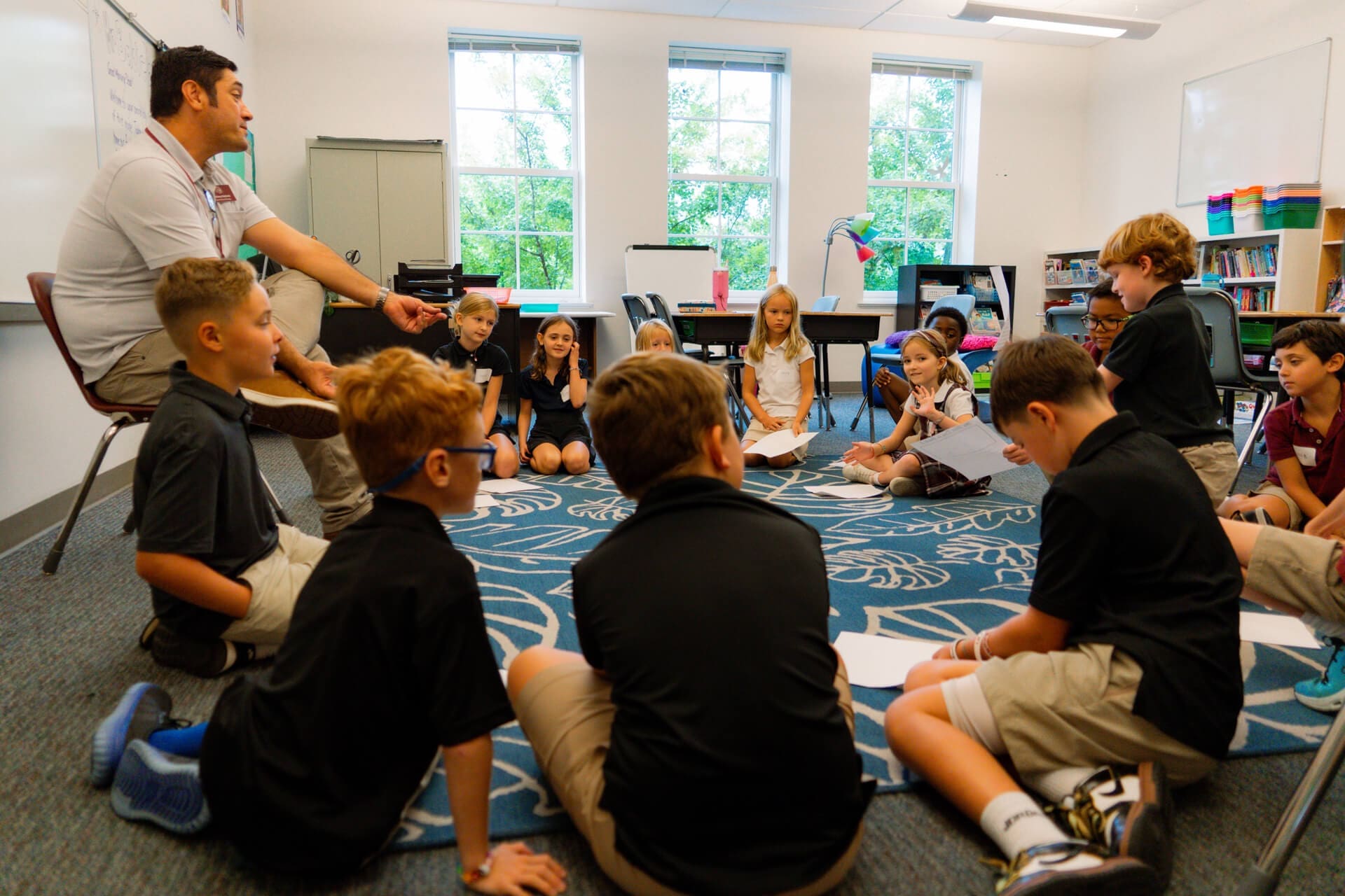 A teacher and a group of elementary school students sit in a circle on a blue patterned rug in a classroom, engaging in a discussion or activity. Desks, shelves, and windows are visible in the background.
