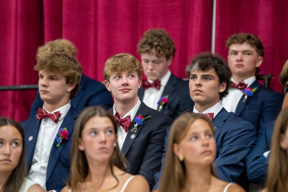 Young men in blue suits and red bow ties sit in rows at an event, possibly a graduation, with a red curtain behind them. Several young women in white outfits are seated in front. Everyone looks attentive.