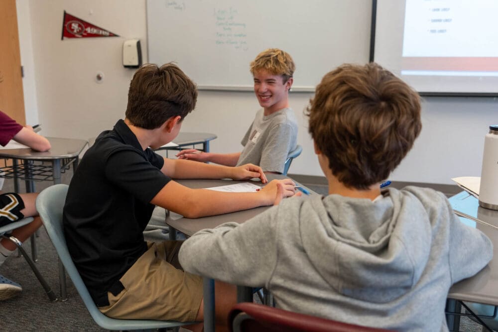 Three teenage boys sit at desks in a classroom, talking and smiling. One boy faces the camera and laughs, while the other two have their backs to the camera. Classroom materials and a whiteboard are visible.