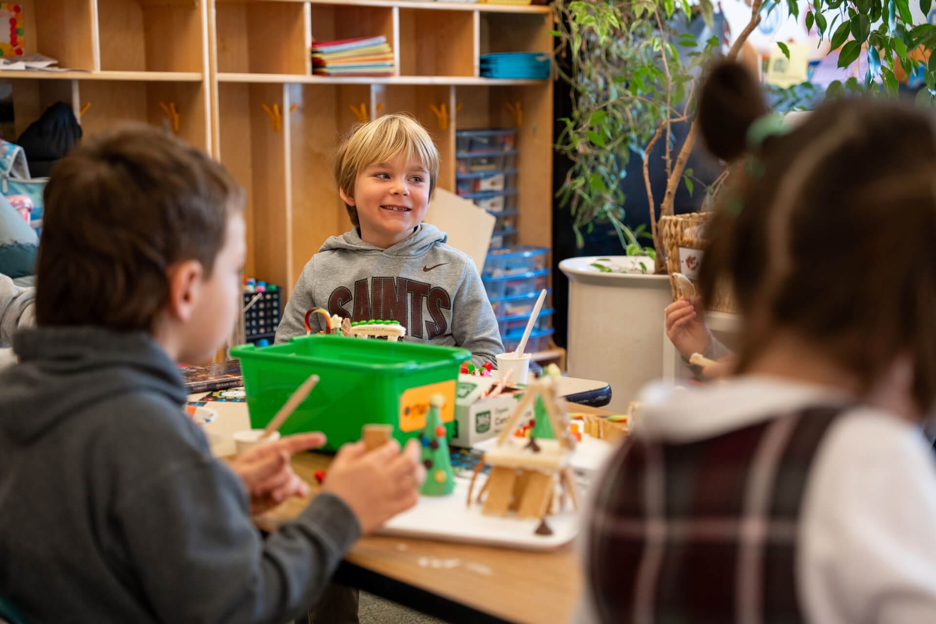 Young children sit around a table in a classroom, engaging in arts and crafts activities. A smiling boy in a gray hoodie is the focus, with colorful supplies and projects visible on the table. Shelves and a plant are in the background.