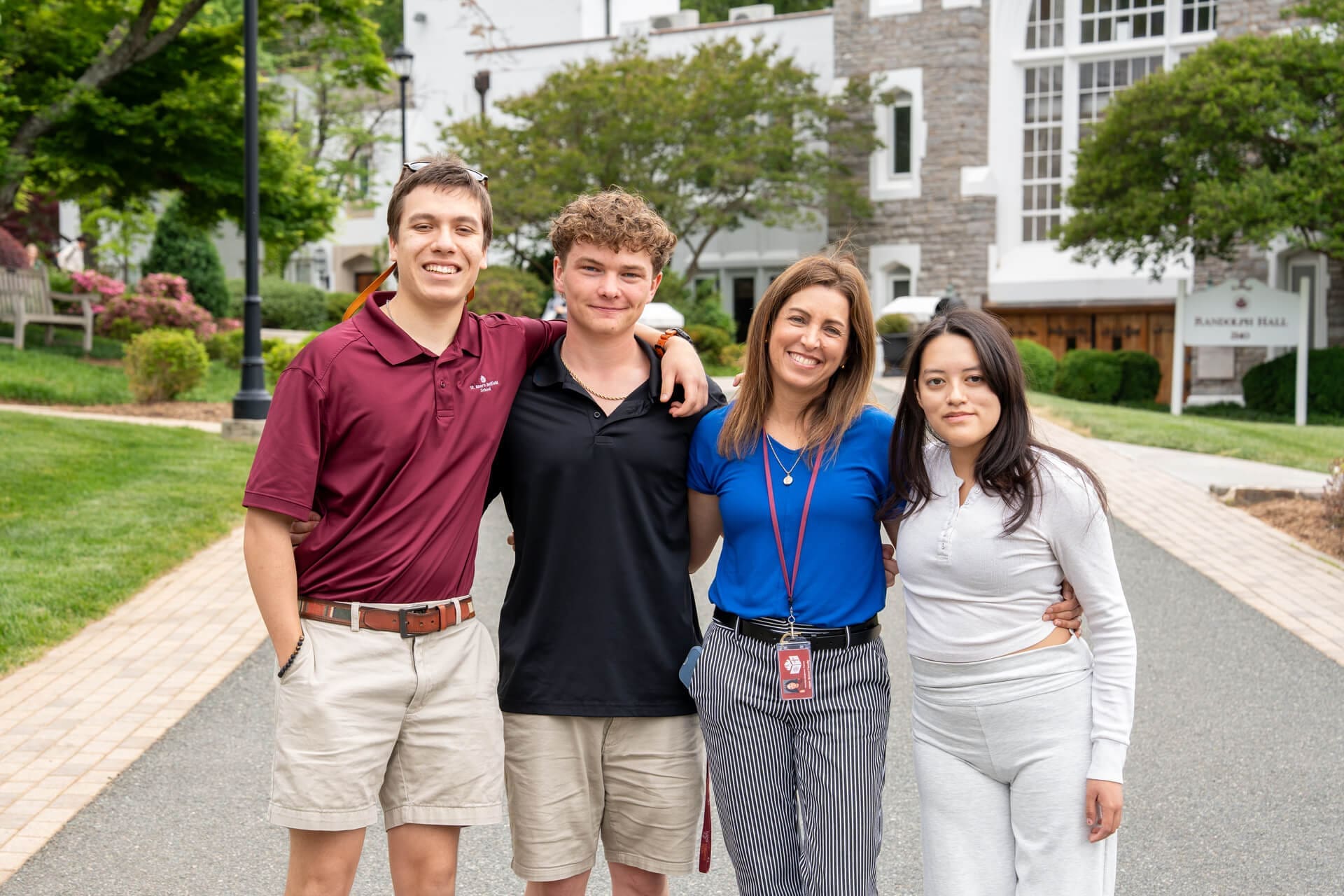 Four people stand outside on a paved path, smiling at the camera. The group includes two young men, a woman in a blue shirt with a lanyard, and a young woman in white. Trees and a building are visible in the background.