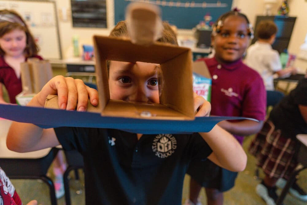A child looks through a cardboard tube while holding a craft project in a school classroom. Other children stand nearby, with a chalkboard and desks in the background.