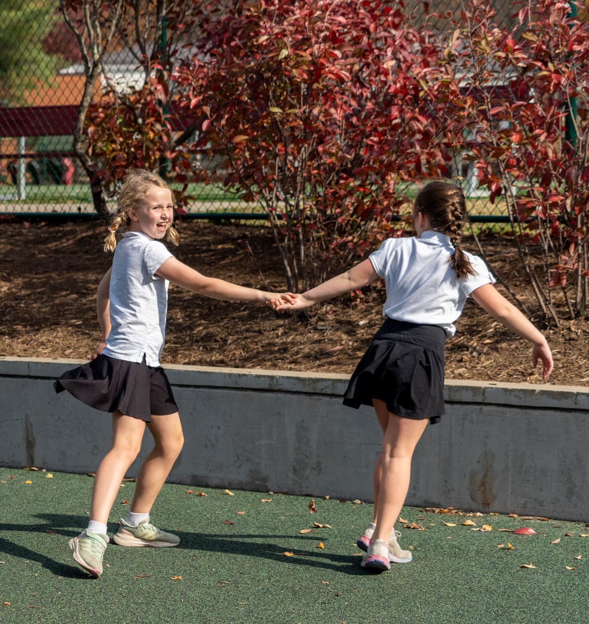 Two young girls wearing white shirts and black skirts hold hands while spinning and playing outside on a sunny day, with red-leafed bushes and a fence in the background.