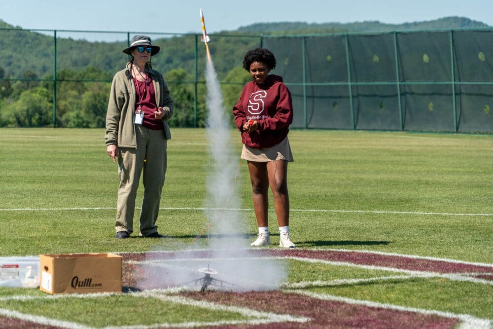 Two people stand on a grassy field, watching a model rocket launch with smoke rising. One person wears a hat and sunglasses; the other is in a maroon sweatshirt with an S. A cardboard box sits nearby.
