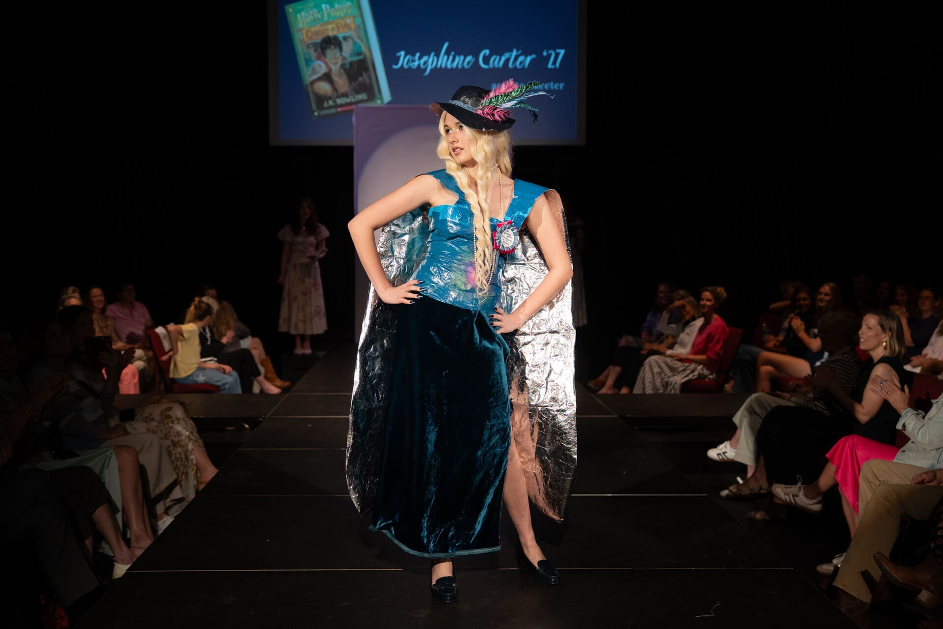 A model in a blue corset dress, large hat with flowers, and dramatic cape poses confidently on a runway, with an audience seated on both sides and a projected sign reading “Josephine Carter ’27” in the background.