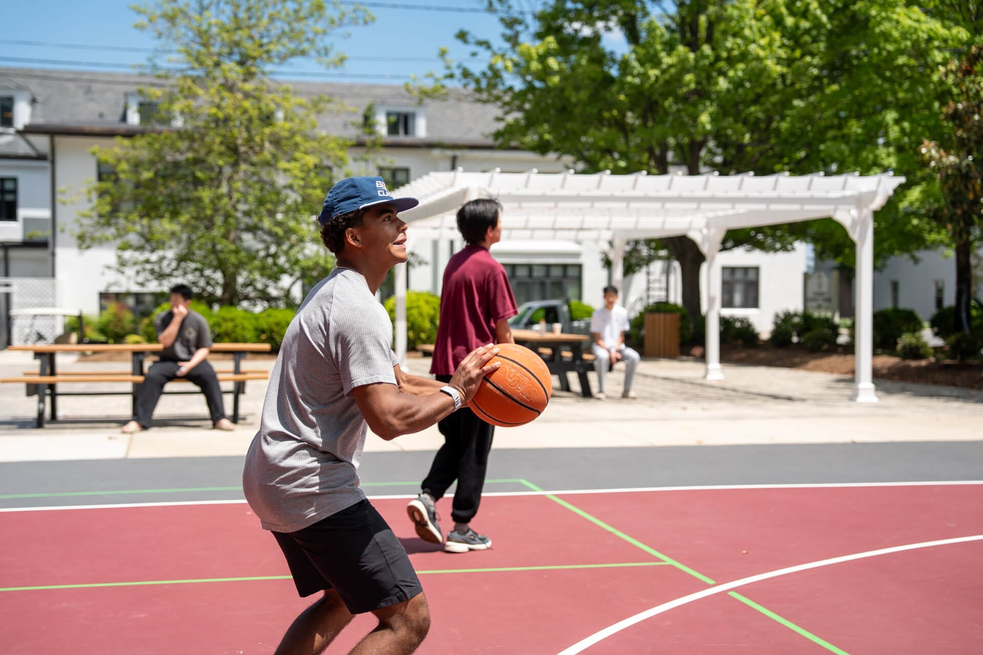 A young man prepares to shoot a basketball on an outdoor court, while another person walks nearby. Two people sit at a picnic table in the background under a pergola, with trees and buildings visible.