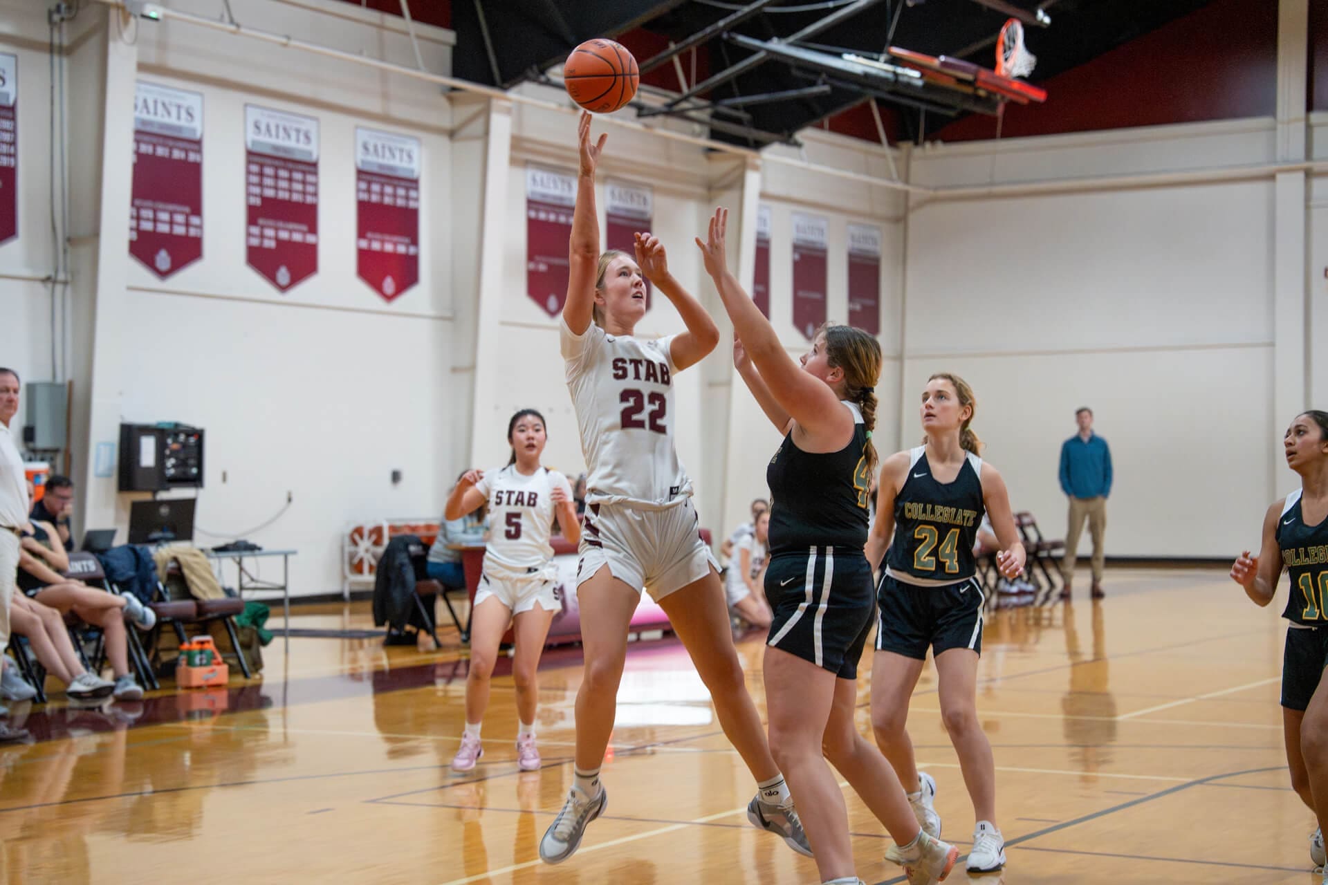 A female basketball player in a white uniform, number 22, jumps to shoot the ball as opposing players in black uniforms attempt to block her on an indoor basketball court. Spectators and banners are visible in the background.