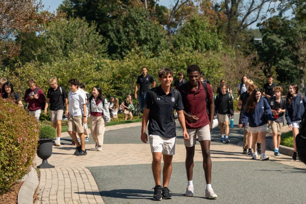A group of high school students in uniforms walk outside on a sunny day, talking and smiling as they follow a curved path lined with greenery and trees.