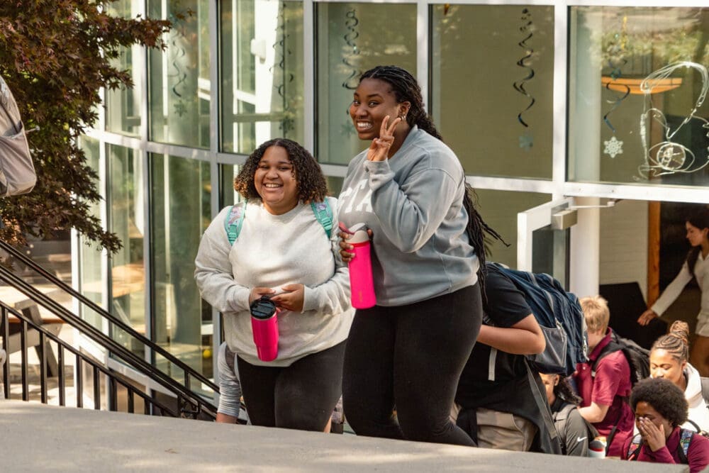 Two students smile while walking up outdoor steps, both holding pink water bottles and wearing backpacks. Other students are seen in the background near a glass building decorated with hanging ornaments.