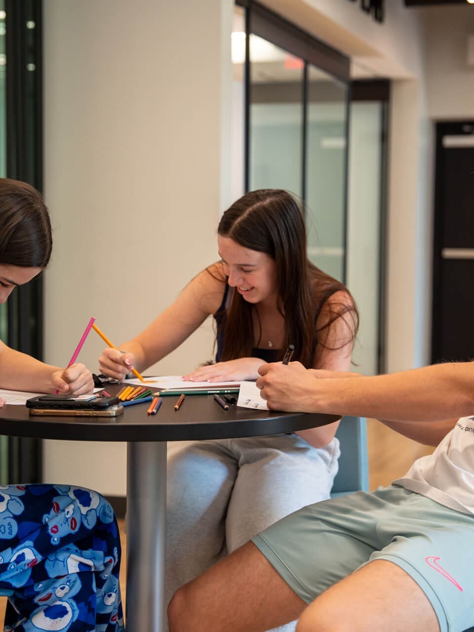 Three young people sit around a small table, smiling and drawing with colored pencils on paper. Art supplies are scattered on the table, and they appear to be working together in a bright indoor space.