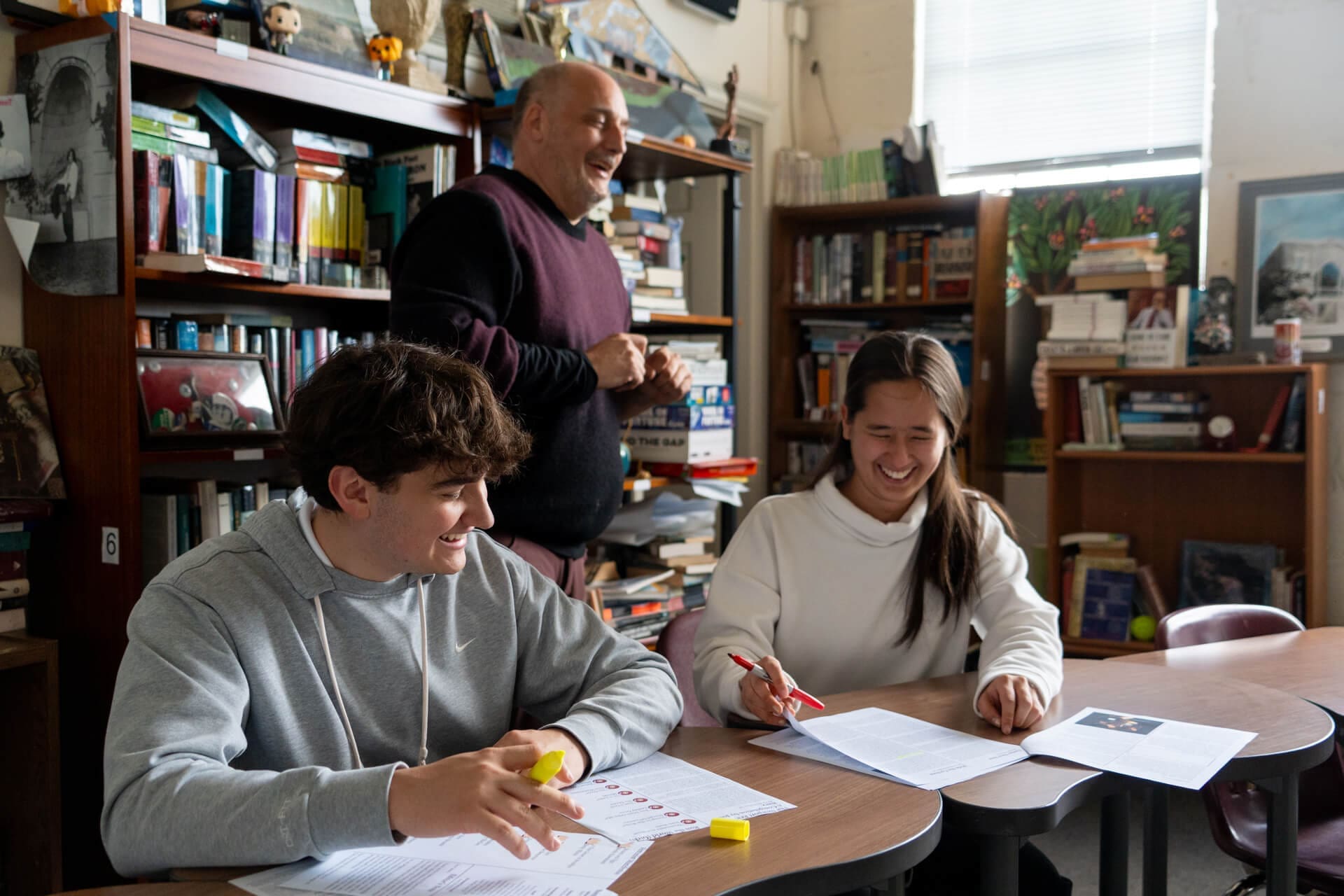 Two students sit at a desk in a school classroom, smiling and working on papers with highlighters, while an older man stands behind them laughing. Bookshelves filled with books and papers are in the background, creating a cheerful atmosphere.