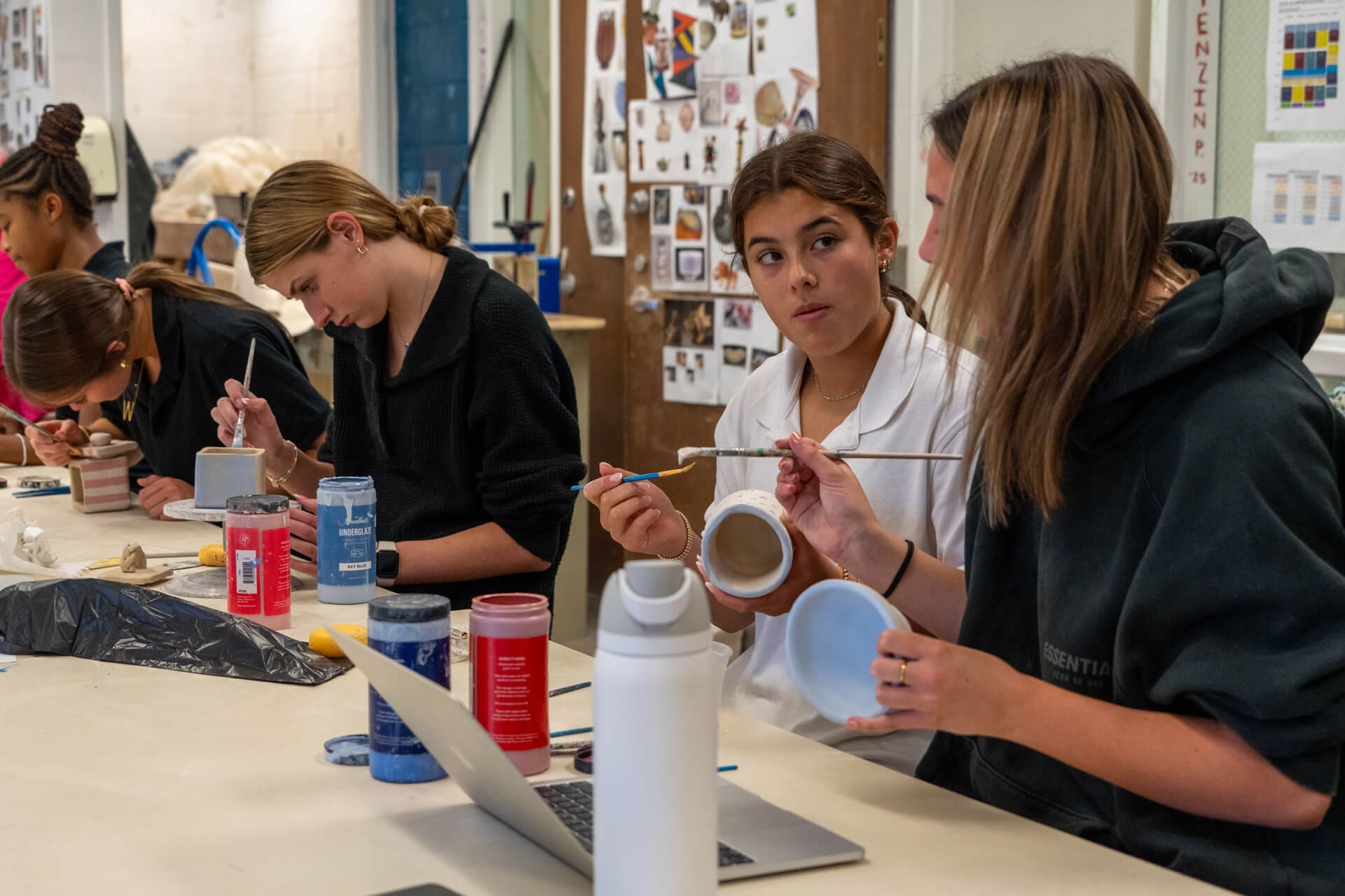 Several young women sit at a table painting pottery in an art classroom, with various paint bottles, brushes, water bottles, and a laptop on the table. A bulletin board with artwork is in the background.