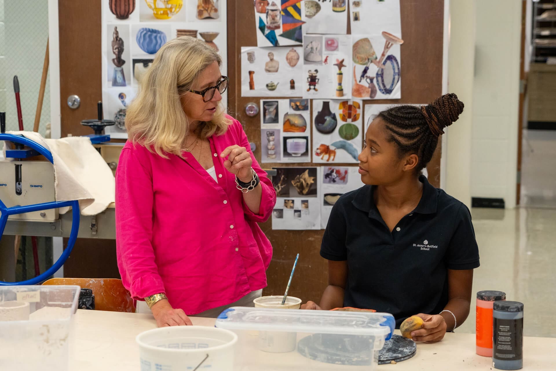 Two women in an art classroom talk while sitting at a table with pottery supplies. One woman wears a bright pink shirt and glasses, while the other wears a black polo. Artworks are displayed on the wall behind them.